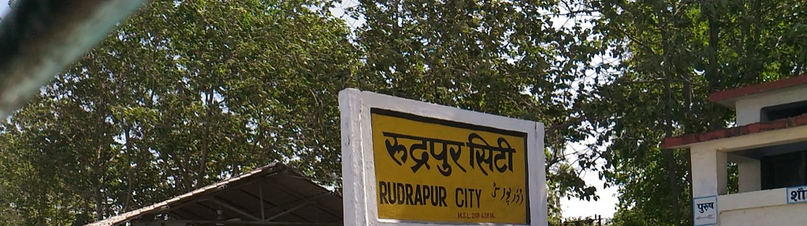 रुद्रपुर सिटी रेलवे स्टेशन का पीला नामपट्ट, जिस पर हिंदी, अंग्रेज़ी और उर्दू में स्टेशन का नाम लिखा है।